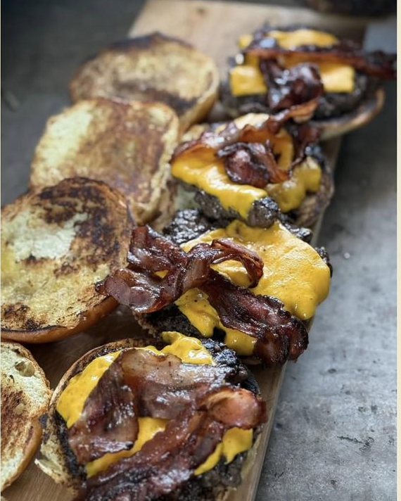 Hamburguesas abiertas con queso derretido y tocino alineadas sobre una tabla de madera.